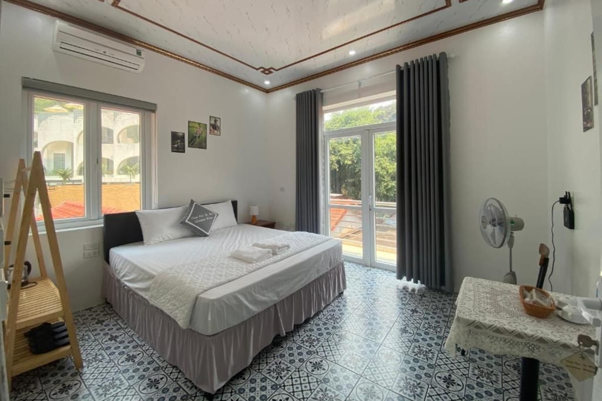 A hotel room with a bed, a fan, a table with a basket, and a window with curtains.