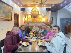 A group of people dining at a table with a chandelier and a speaker in the background. - Tam Coc Harmony Homestay (Nam Hoa Lư)