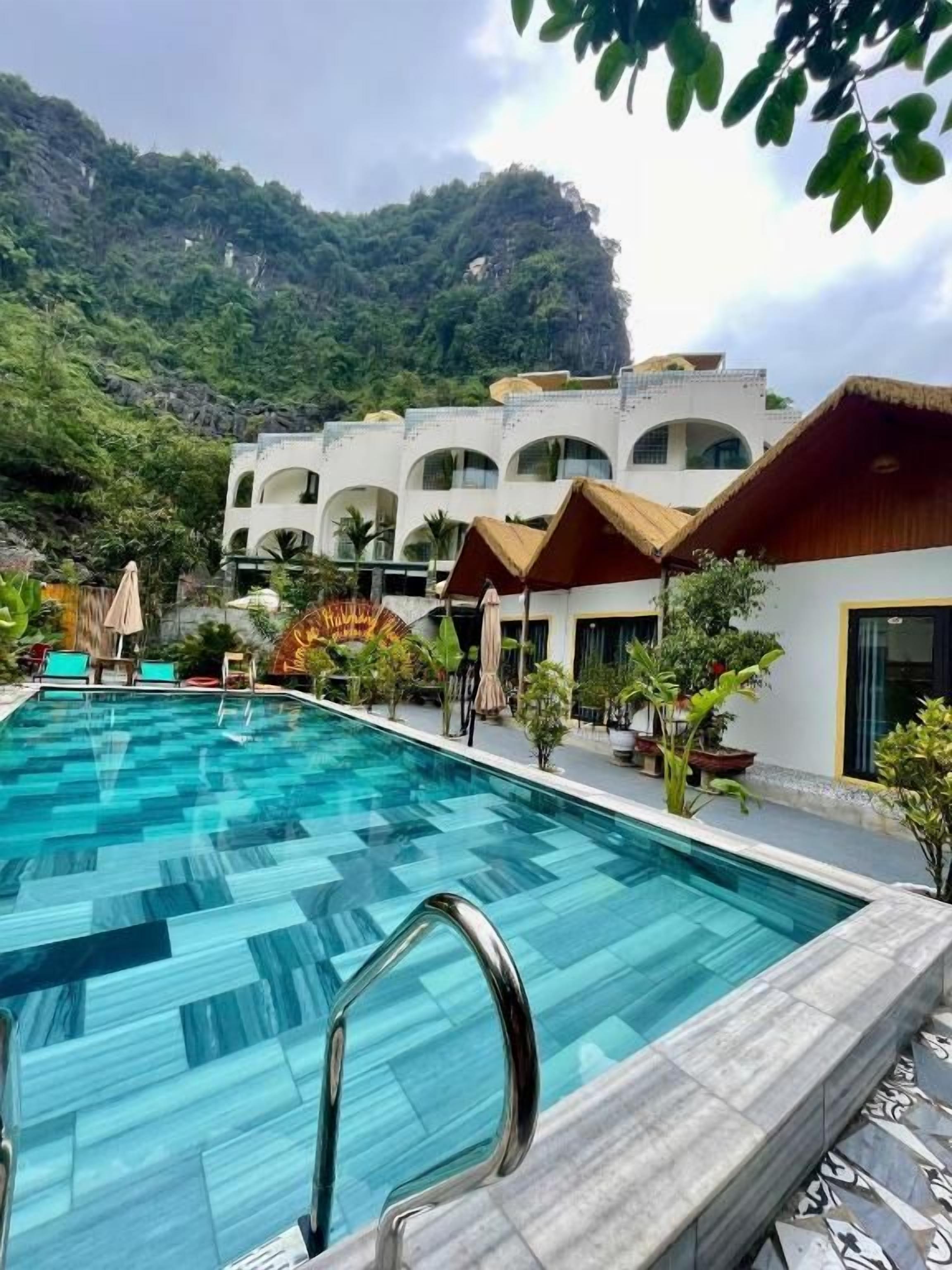 Una piscina con un patrón de agua azul cristalina, rodeada por un edificio con varios balcones y montañas al fondo.
