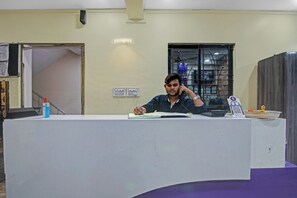 A man sitting at a desk in a room with a window, a door, and a wall-mounted light fixture. - Hotel O Gkumar Restro (Nagpur)