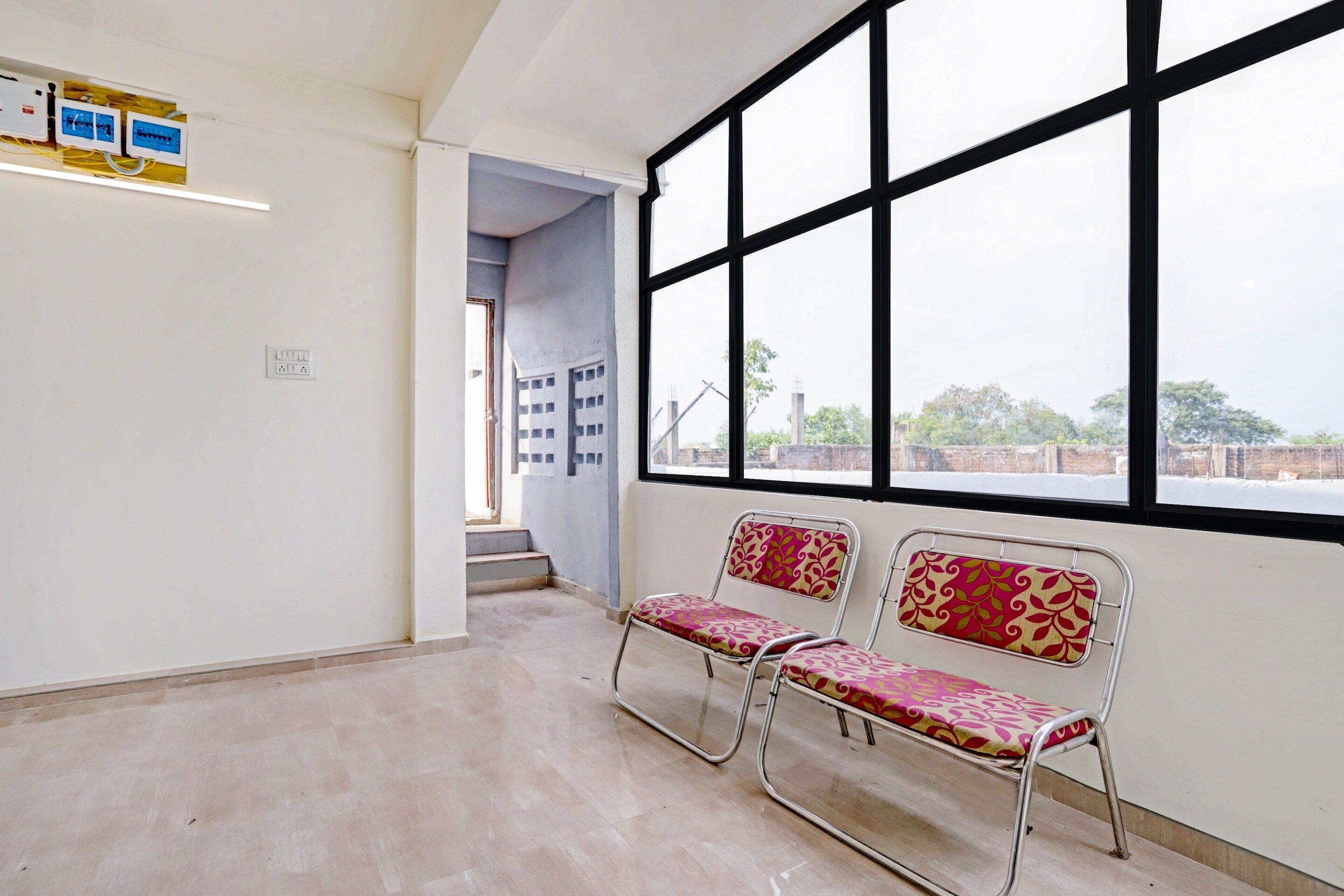 A waiting area with two metal chairs, a window with black frames, and a wall-mounted electrical panel.