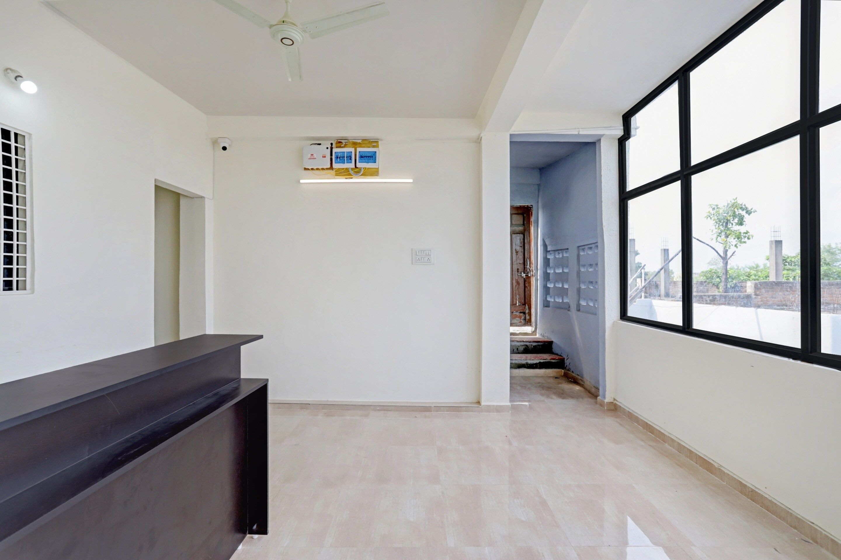 A hotel reception area with a black reception desk, a ceiling fan, a security camera, and a window with a view of the outside.
