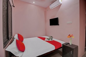 Una habitación de hotel con una cama, una mesita de noche con un hervidor, un jarrón con flores y un televisor en la pared.