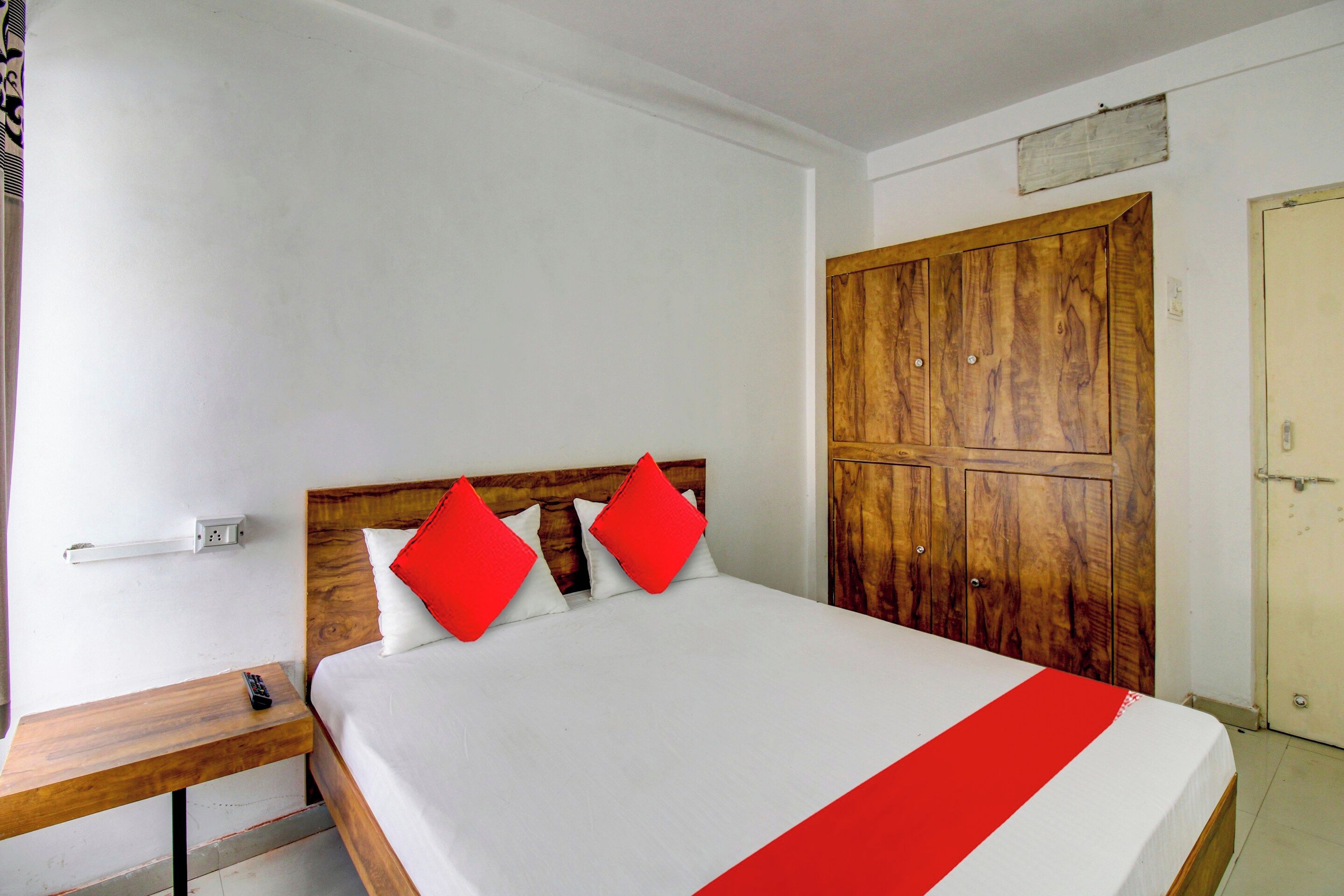 Un letto singolo con poggiatesta in legno, due cuscini, un comodino con telecomando e un armadio con anta in legno.