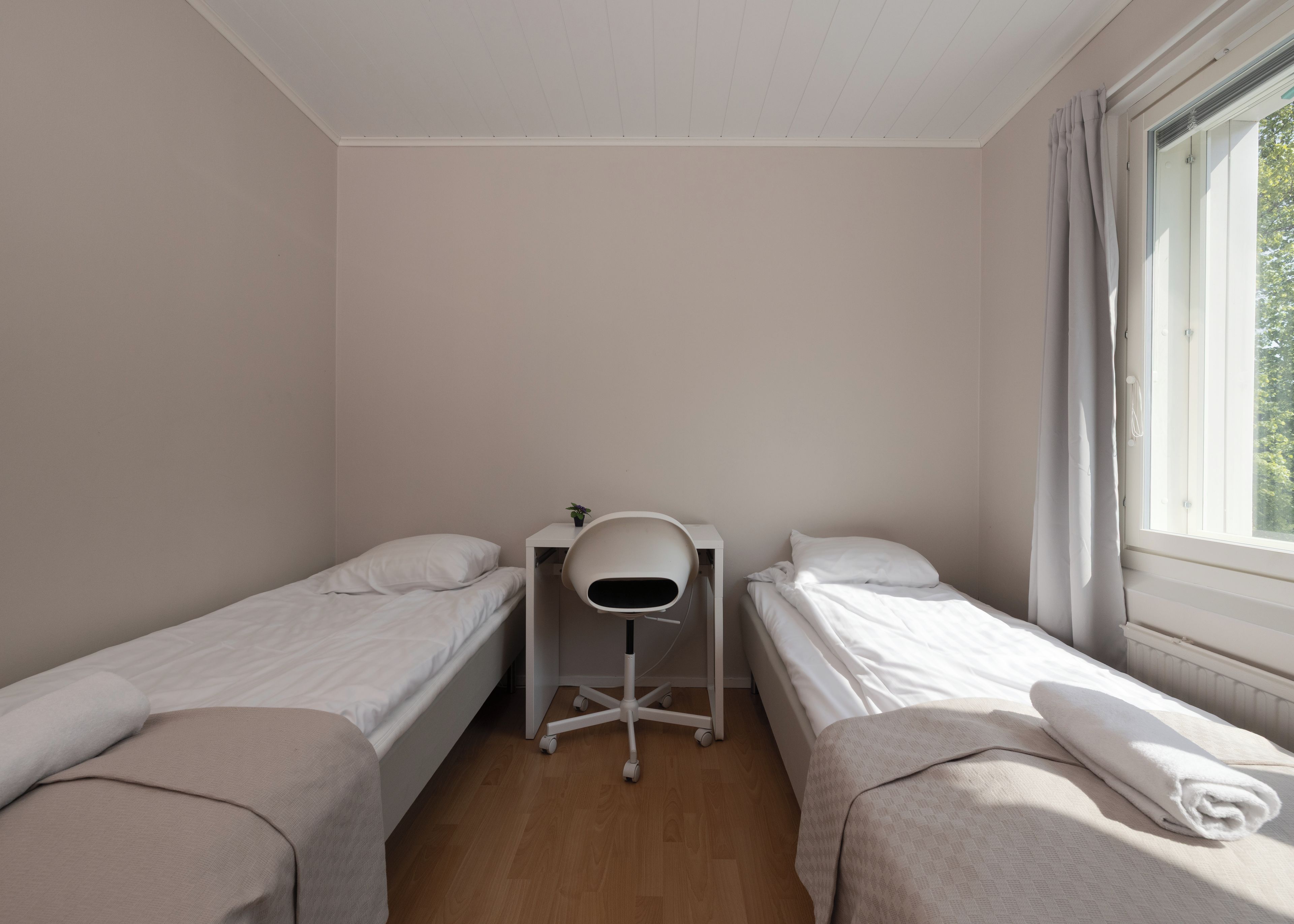 A small, single-room accommodation with two beds, a desk, and a chair.