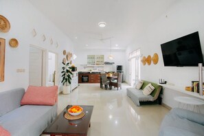 A modern living room with a sofa, a coffee table with a bowl of fruit, a dining area with chairs, a kitchen in the background, and a flat-screen TV mounted on the wall. - House in the Bush (Badung)