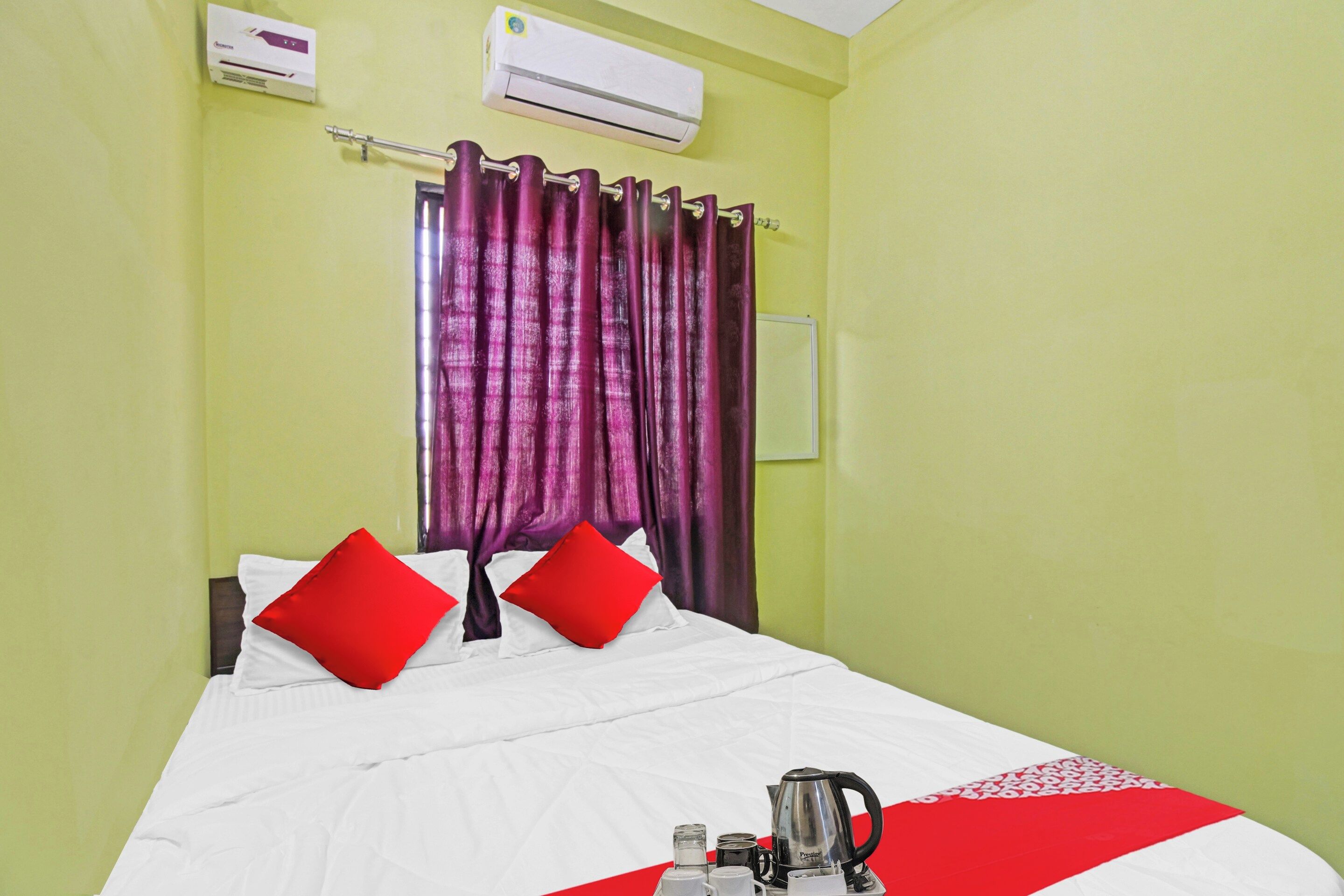 A hotel room with a bed, red pillows, a white bedspread, a red and white striped area rug, a coffee maker, and a white air conditioning unit on the wall.