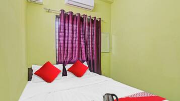 A hotel room with a bed, red pillows, a white bedspread, a red and white striped area rug, a coffee maker, and a white air conditioning unit on the wall.