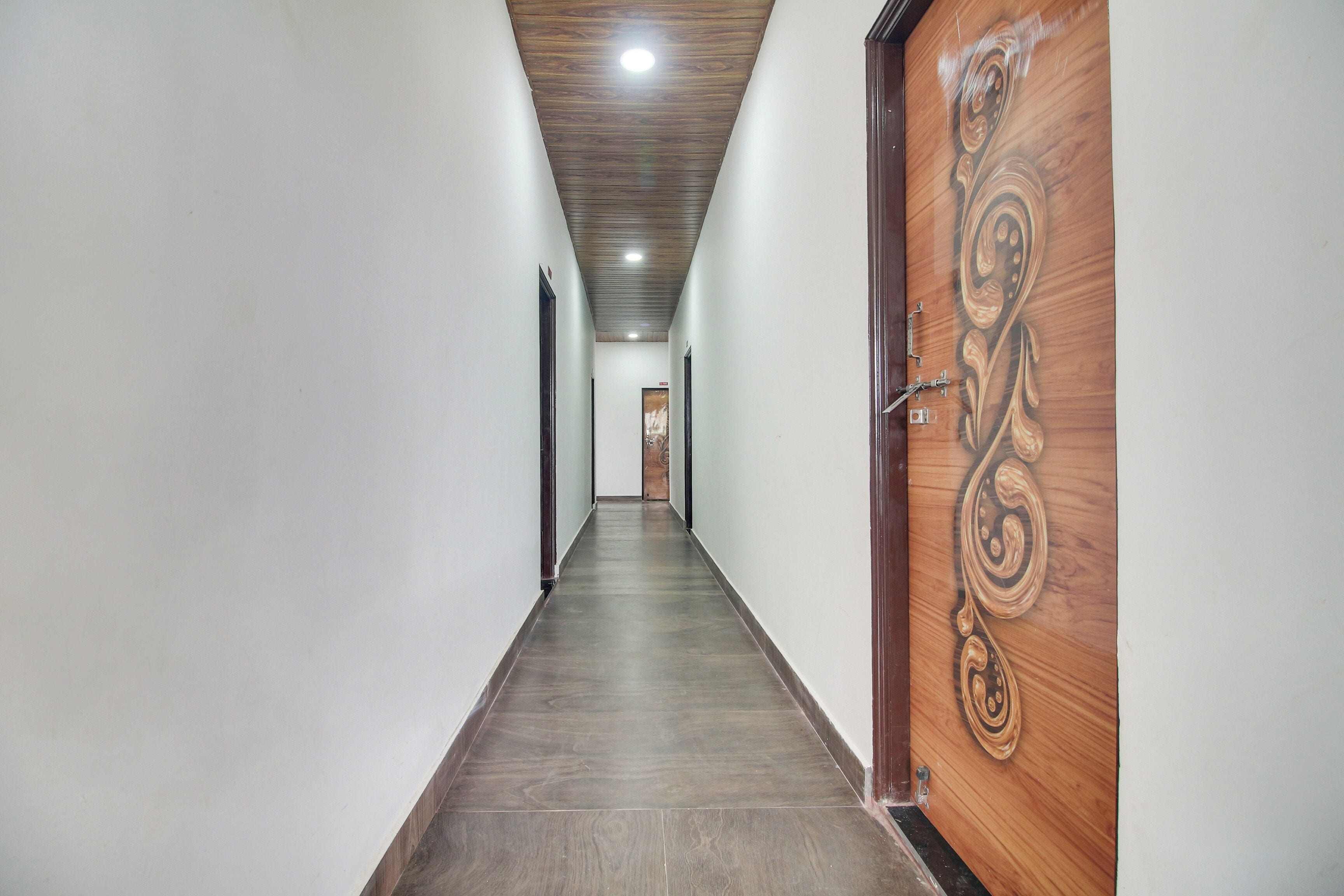 A long, narrow hallway with a wooden door on the right, a light-colored wall, and a dark floor.