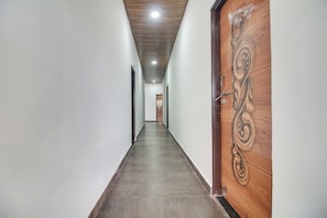 Um corredor comprido e estreito com uma porta de madeira no lado direito, parede de cor clara e chão escuro.