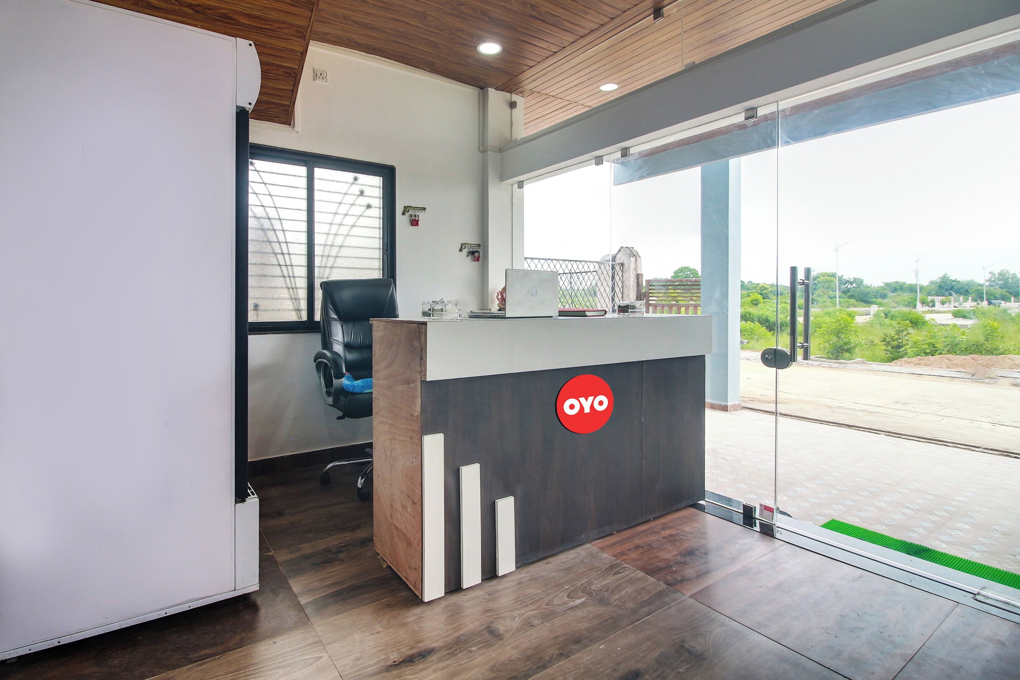 Un área de recepción de oficina con un escritorio de madera, una silla de oficina negra, una computadora portátil y una puerta de vidrio que da al exterior.