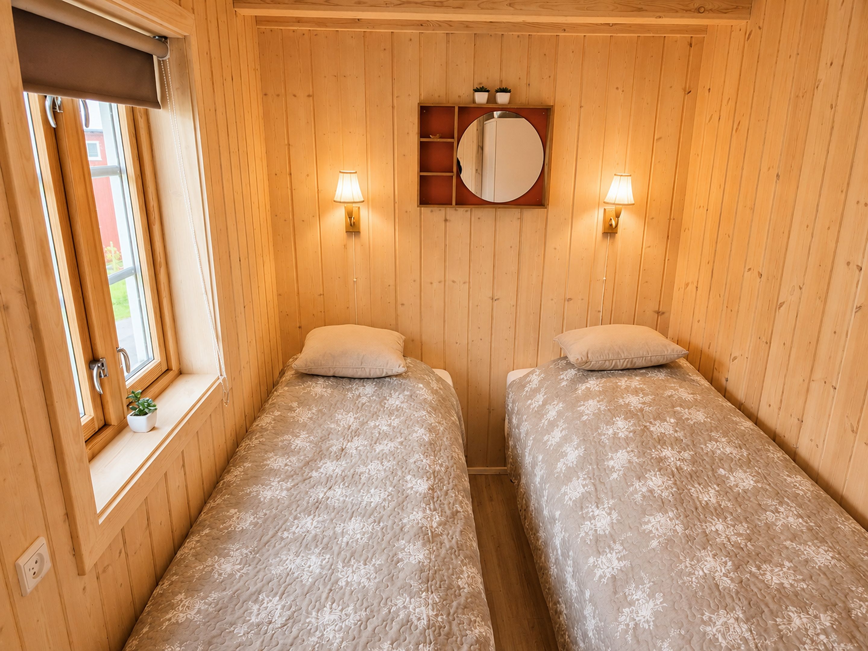 A room with two beds, a window with a view, a mirror, and wall-mounted lights.
