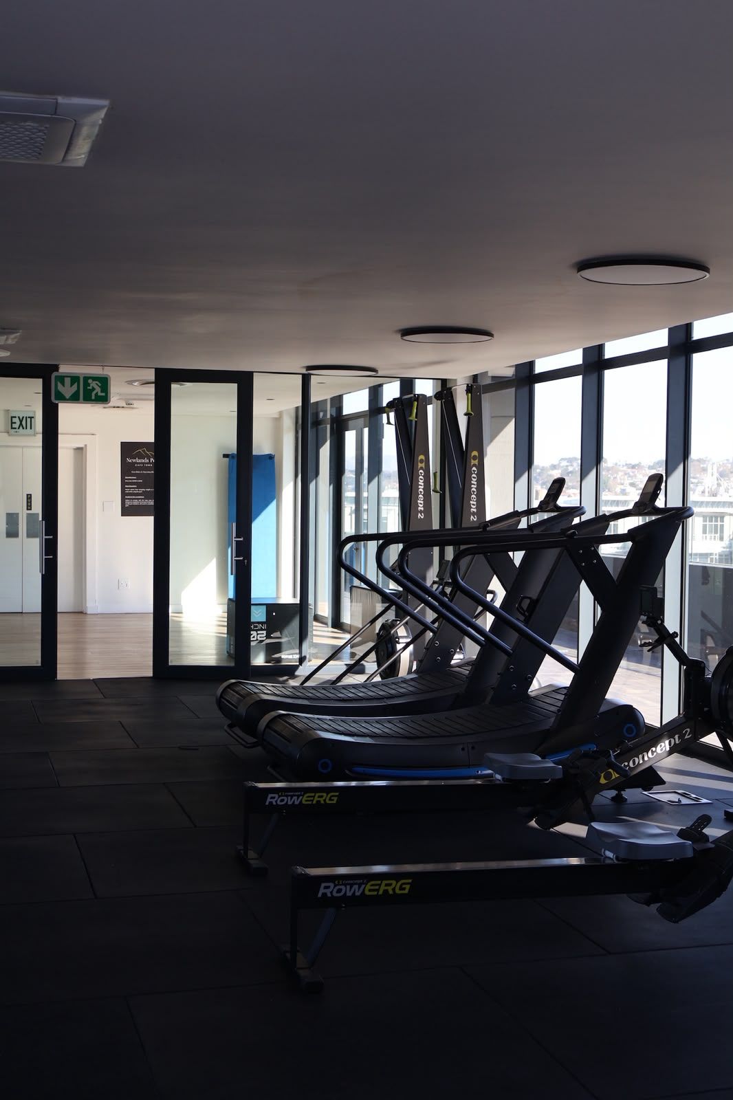 Ein Fitnessstudio mit Trainingsgeräten, darunter Laufbänder und Kraftgeräte, in einem modernen Gebäude mit großen Fenstern.