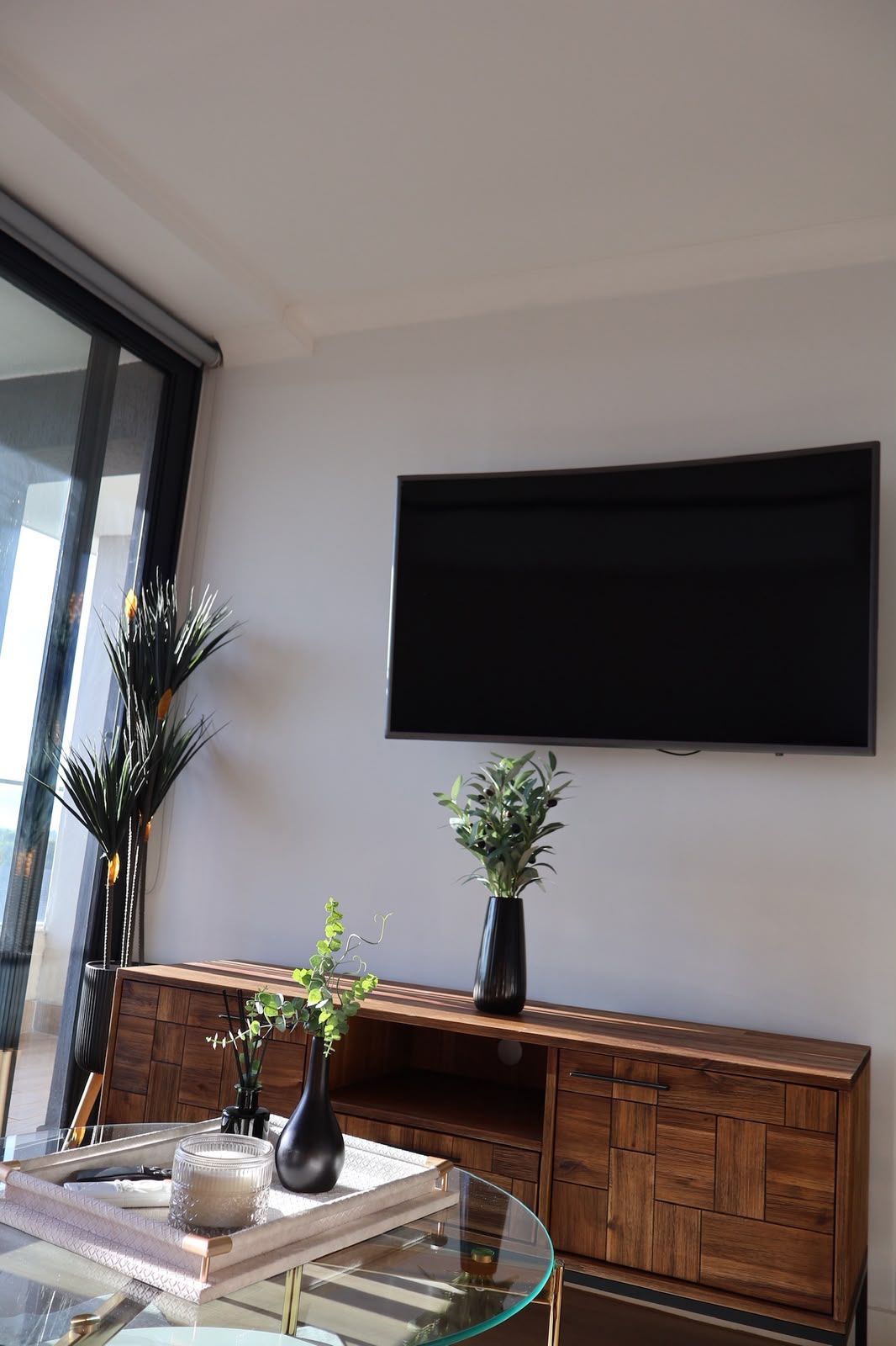 Una sala de estar moderna con un televisor de pantalla plana montado en la pared, un mueble de madera debajo y una mesa de centro de vidrio con objetos decorativos.