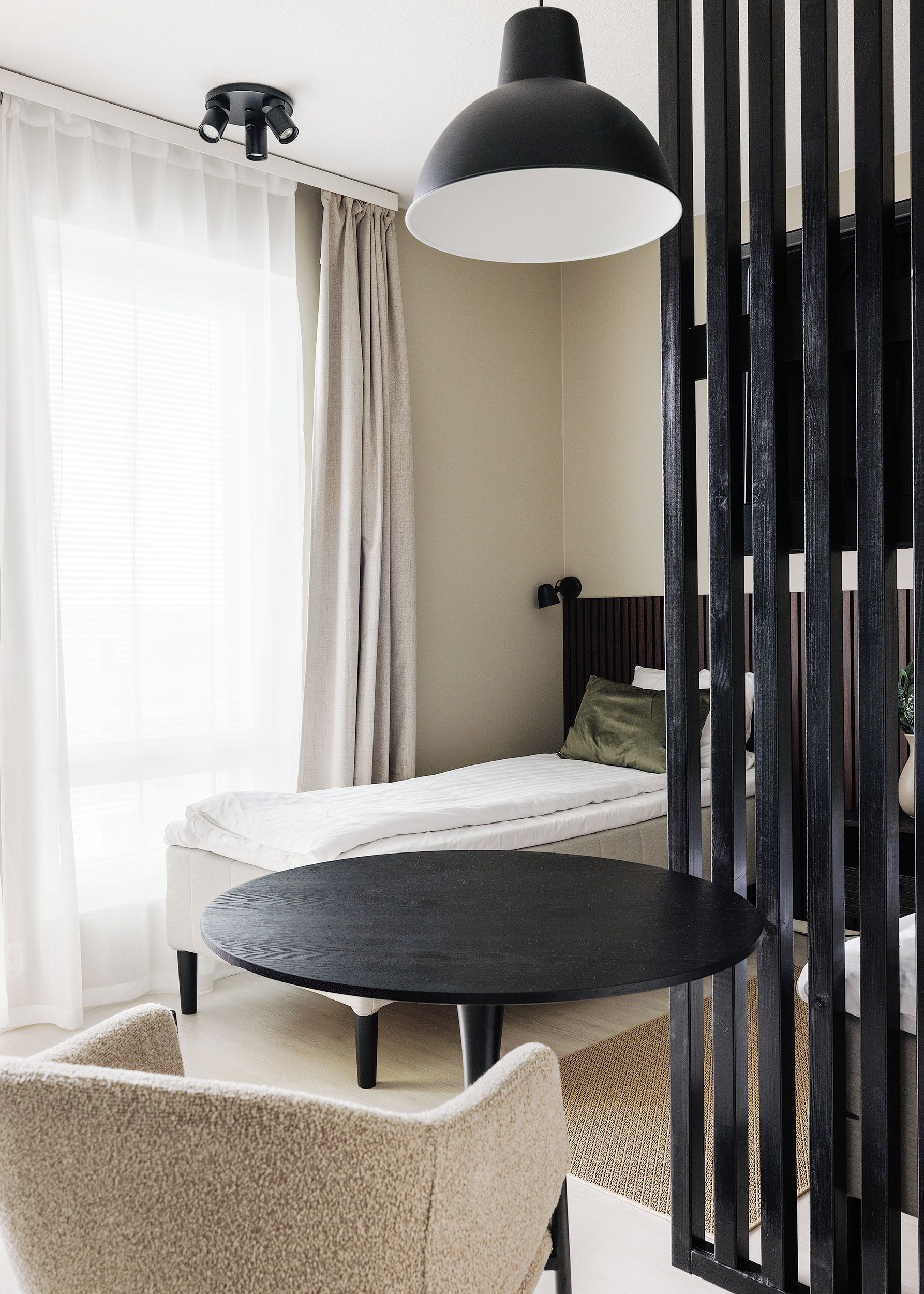 Une chambre moderne avec un lit, une table ronde, une chaise, un luminaire suspendu noir et une fenêtre avec des rideaux.