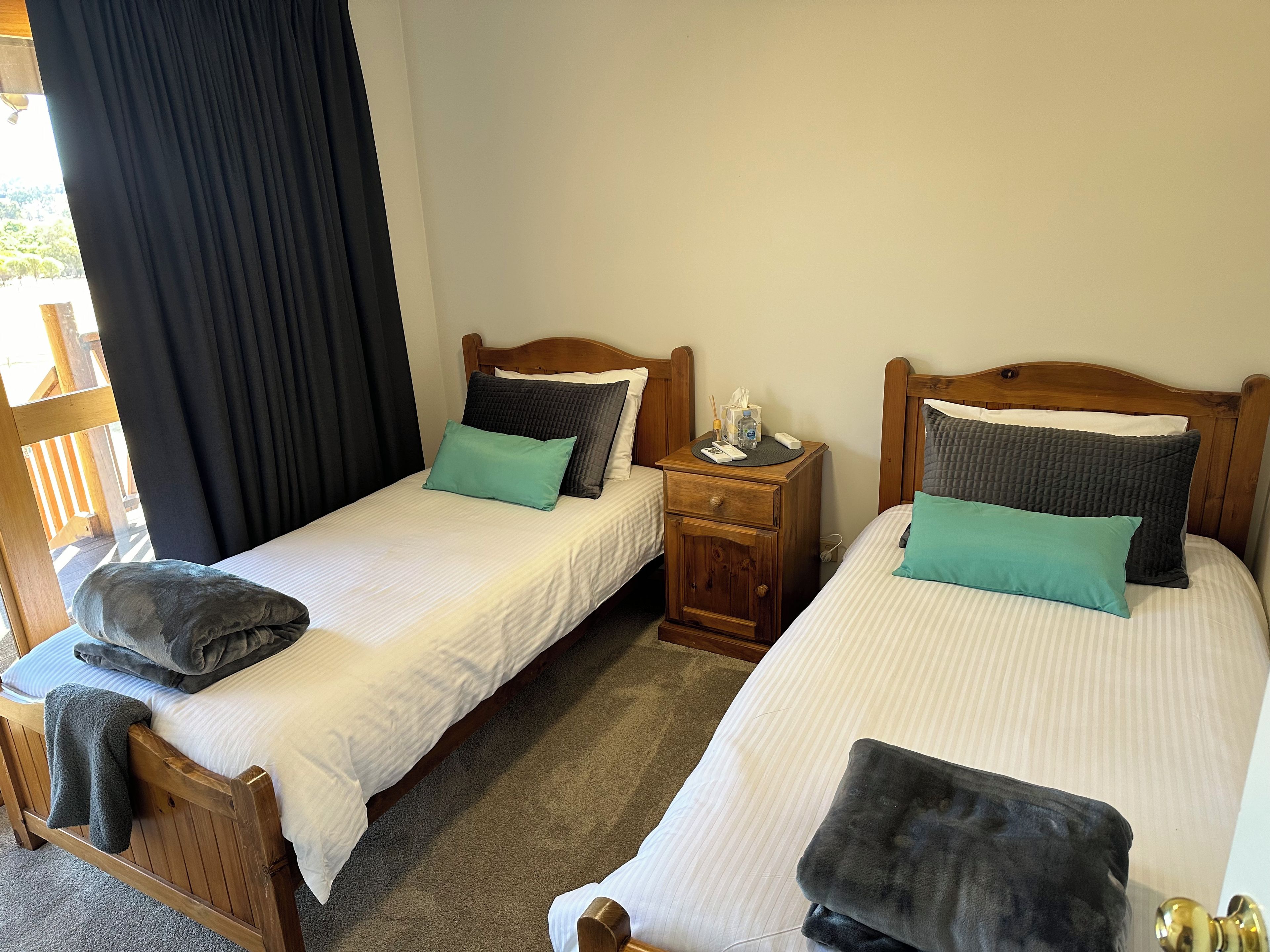 Two single beds with wooden headboards, a nightstand with a lamp, and a window with black curtains.