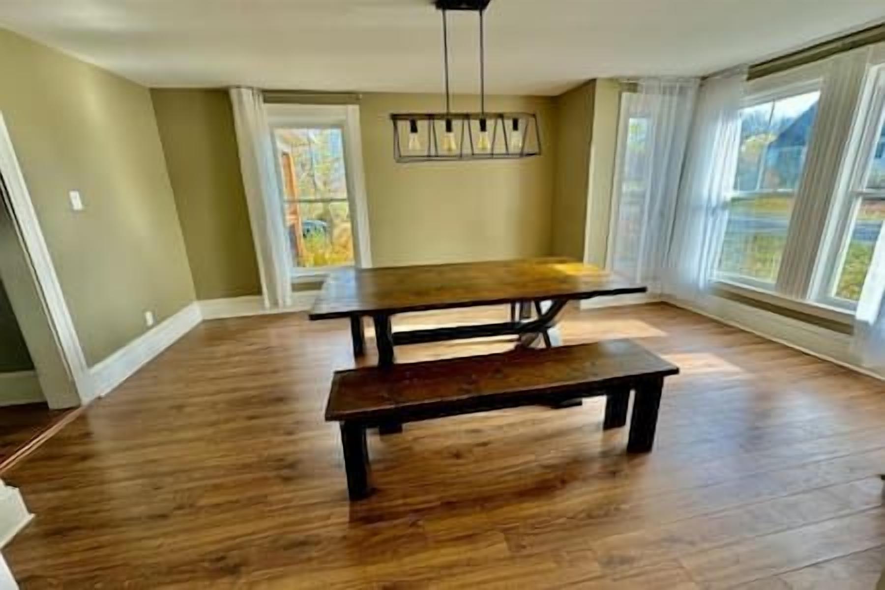 Une pièce avec un plancher en bois, une grande table et un banc. Il y a une fenêtre avec des rideaux et une porte donnant sur l’extérieur.