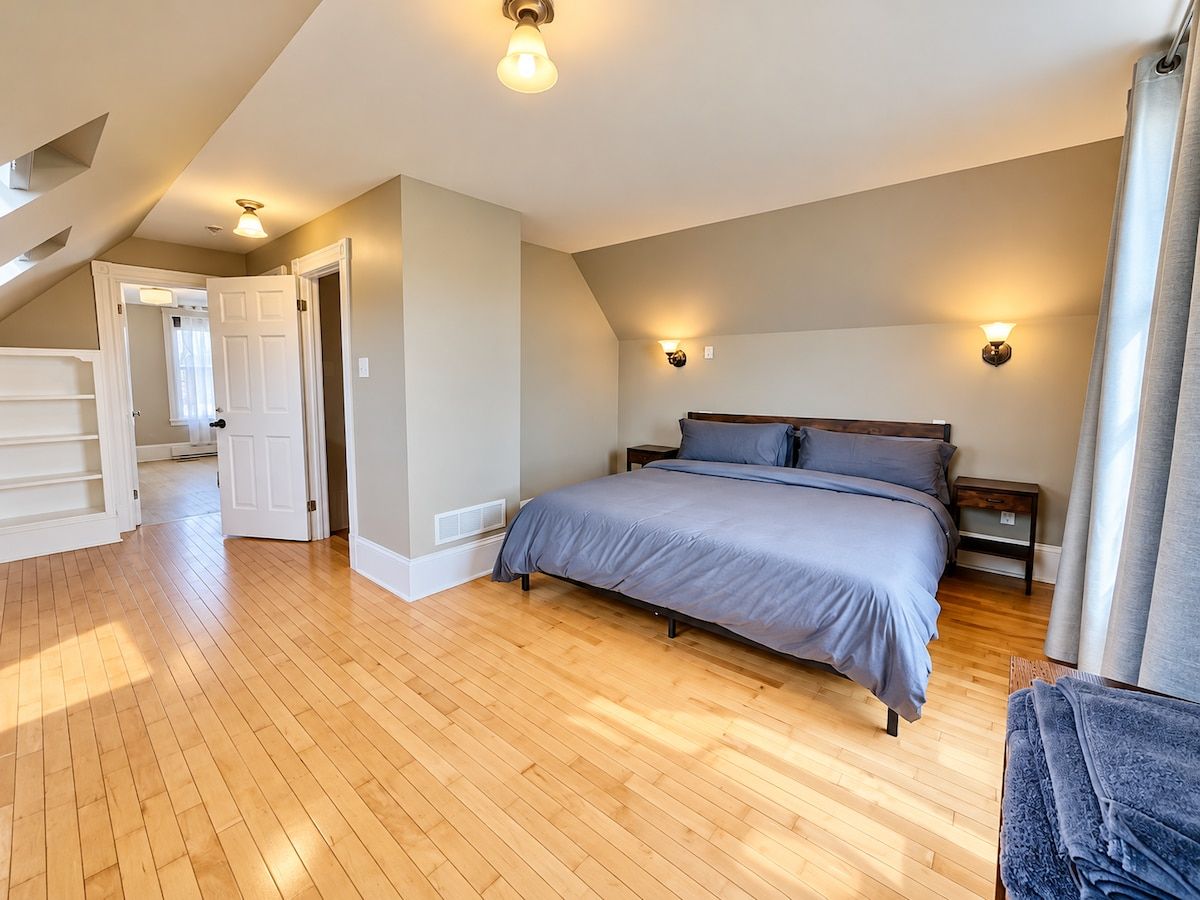 Une chambre avec un plancher en bois, un lit avec une literie grise, deux tables de chevet et un luminaire au plafond.