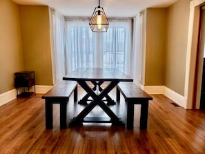 Un coin repas avec une table et des bancs en bois, un luminaire suspendu et une fenêtre ornée de rideaux légers.