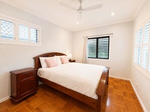 A bedroom with a wooden bed, a nightstand, a dresser, a ceiling fan, and windows with blinds. - Wynnum Bayside Escape 4BR Home with Private Pool (Wynnum)