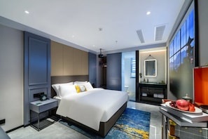 Une chambre d’hôtel moderne avec un grand lit, une table de chevet, un bureau avec une chaise, un grand miroir et une vue sur la ville par la fenêtre.