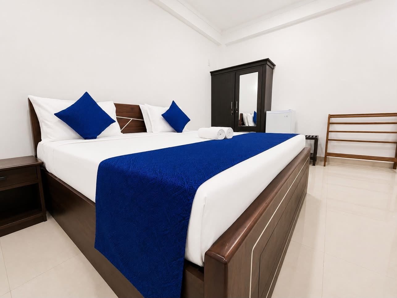 Ein ordentlich bezogenes Bett mit einer blauen Decke, einem dunklen Holz-Kopfstück und einer weißen Wand.
