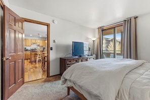 Un dormitorio con cama, televisor sobre un aparador de madera, y vistas a una cocina y balcón a través de un ventanal grande.