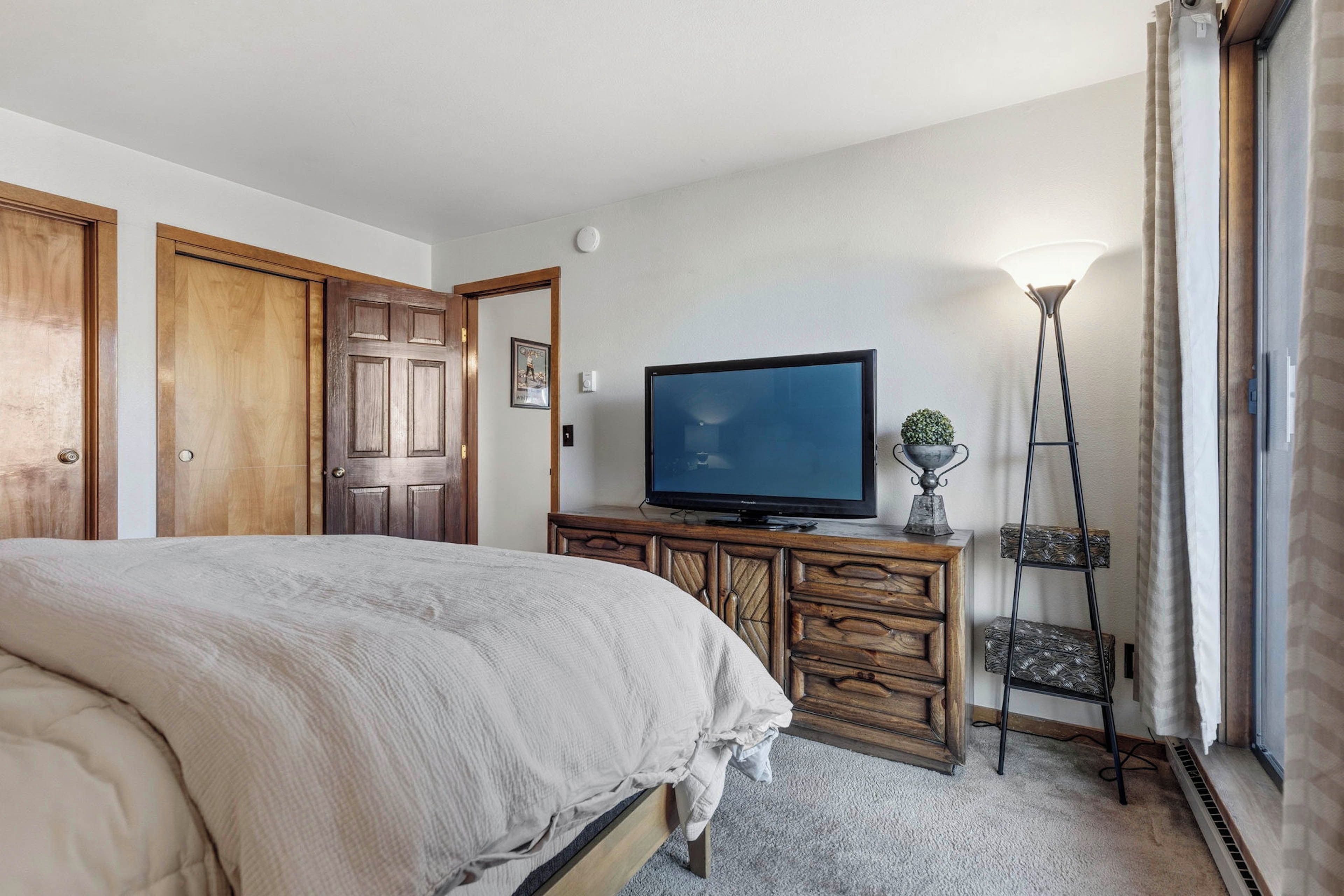 Ein Schlafzimmer mit Bett, Fernseher, Kommode, Lampe und einem Fenster mit Vorhängen.