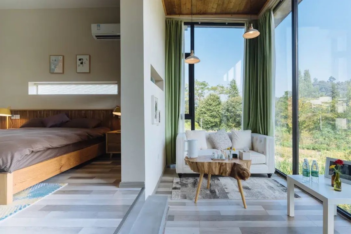 A bedroom with a bed and a view of a green landscape through large windows.