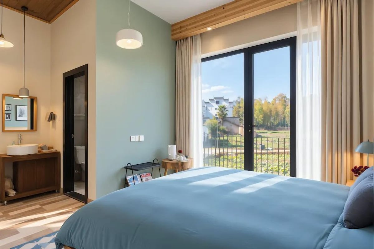 A hotel room with a large bed, a bathroom with a sink, and a view of a park through a large window.