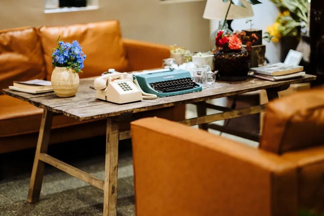 Uma máquina de escrever antiga sobre uma mesa de madeira, um telefone, um vaso com flores azuis e um sofá de couro em um quarto aconchegante.
