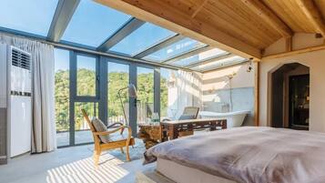 Un dormitorio con una cama grande, una silla de madera, una mesita y vistas al exterior a través de amplias ventanas.