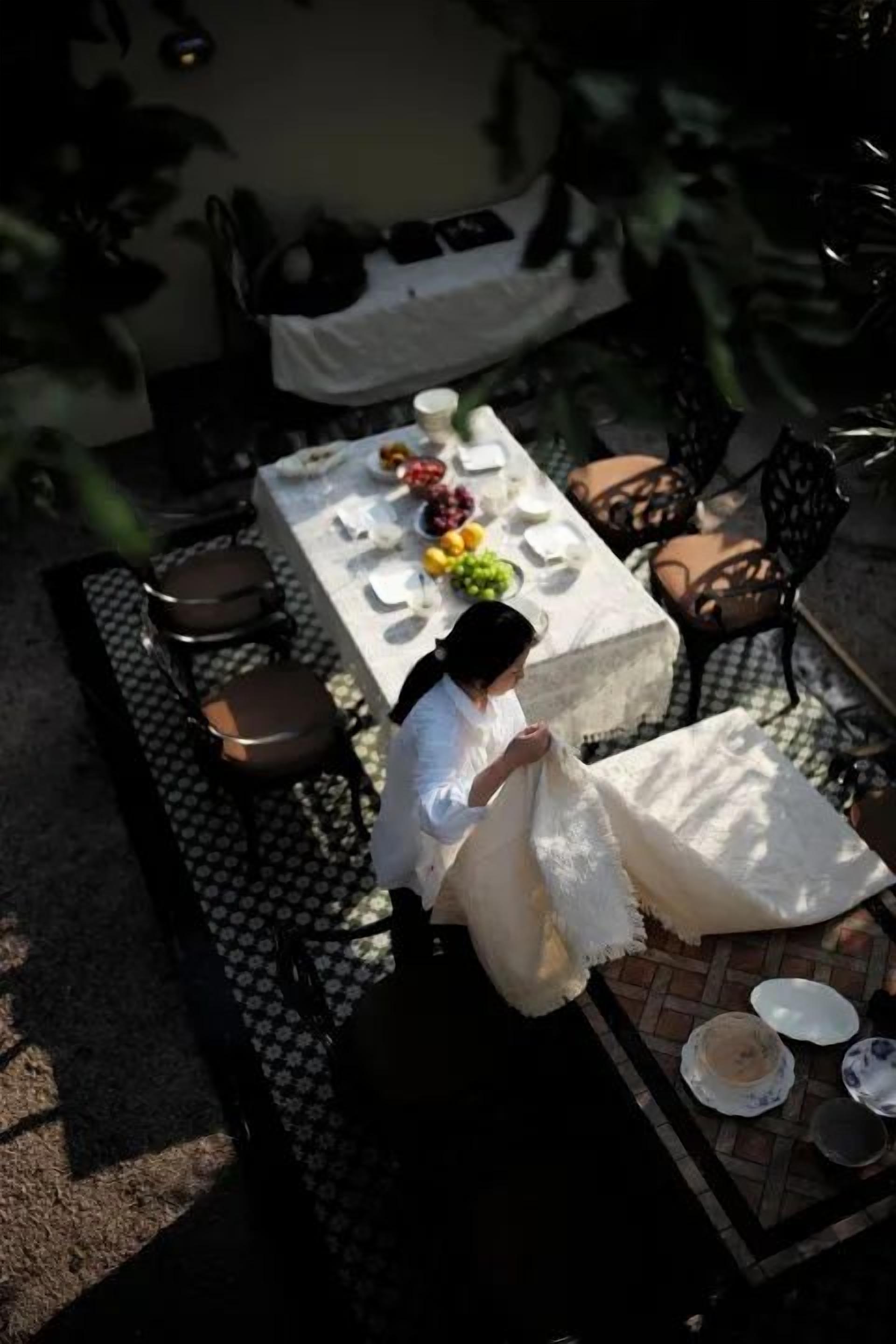 A person seated at an outdoor table with a white tablecloth, surrounded by chairs and a dining set.