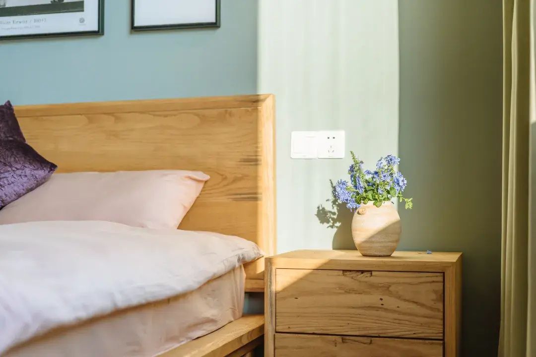 A bedroom with a wooden bed, a nightstand with a vase of flowers, and a wall socket.