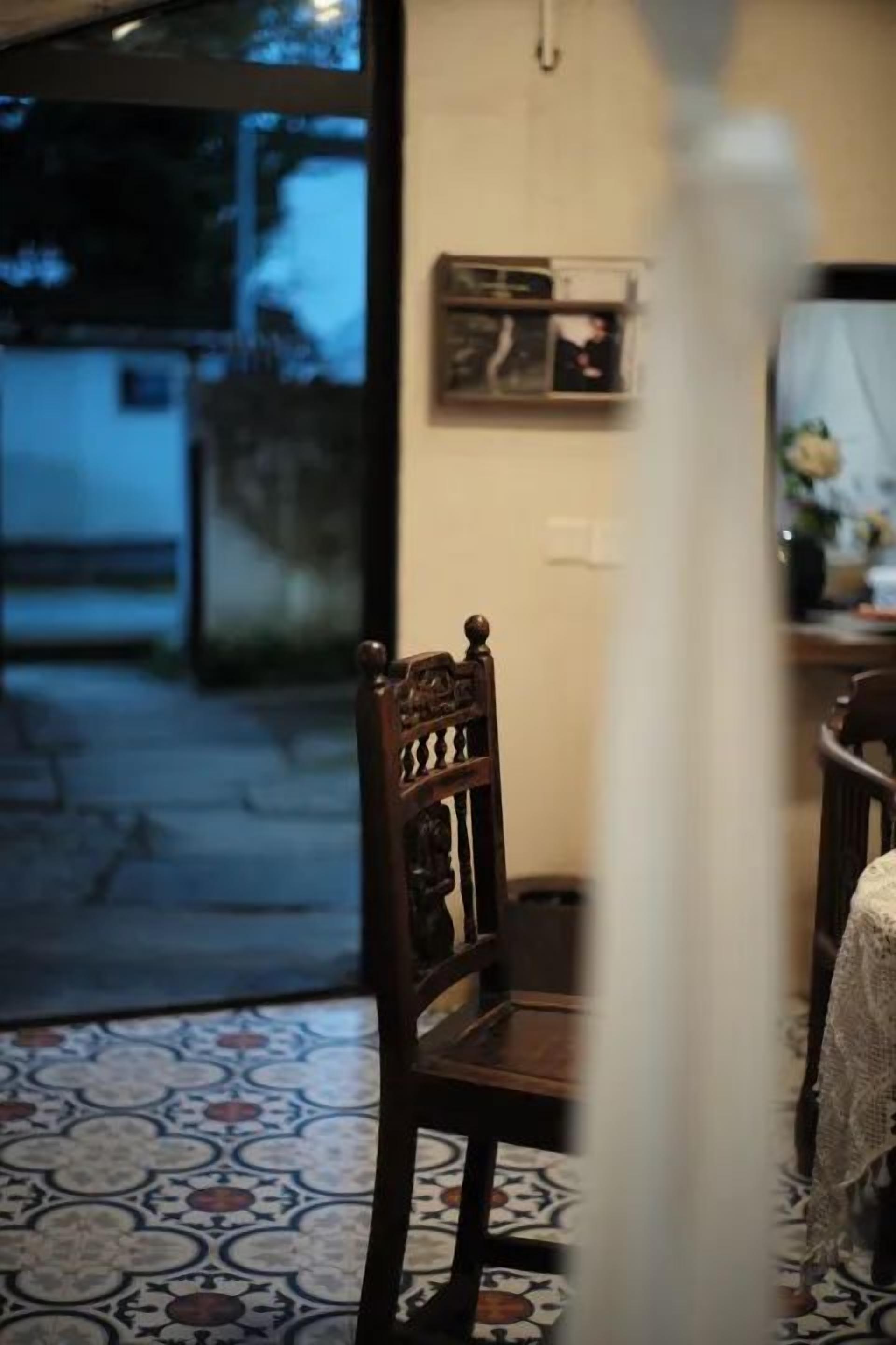 Una habitación con piso estampado, una silla de madera, una mesa con un jarrón y una ventana con vista al exterior.