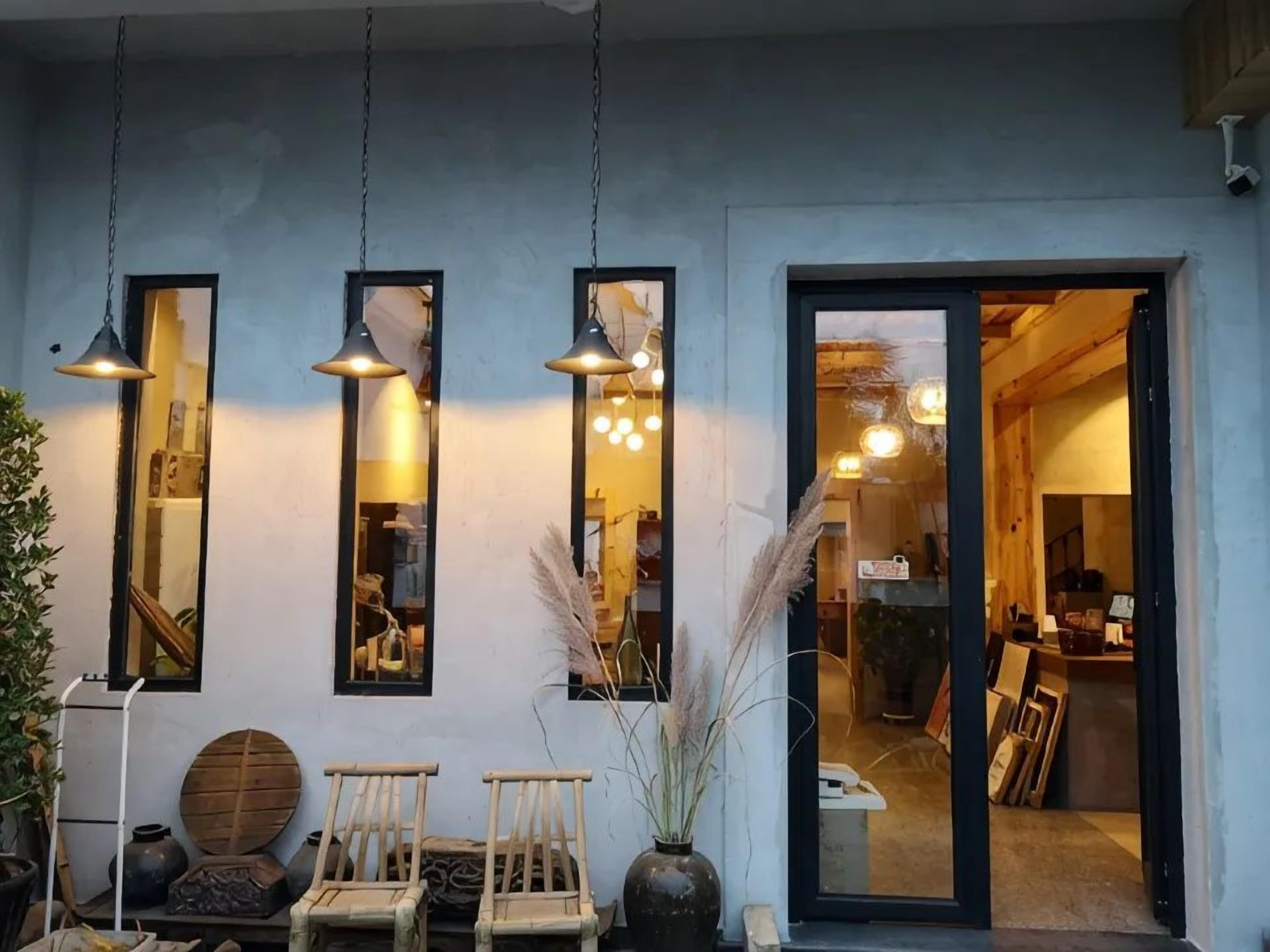 A storefront with hanging pendant lights, a wooden chair, a round table, and a vase with dried plants.