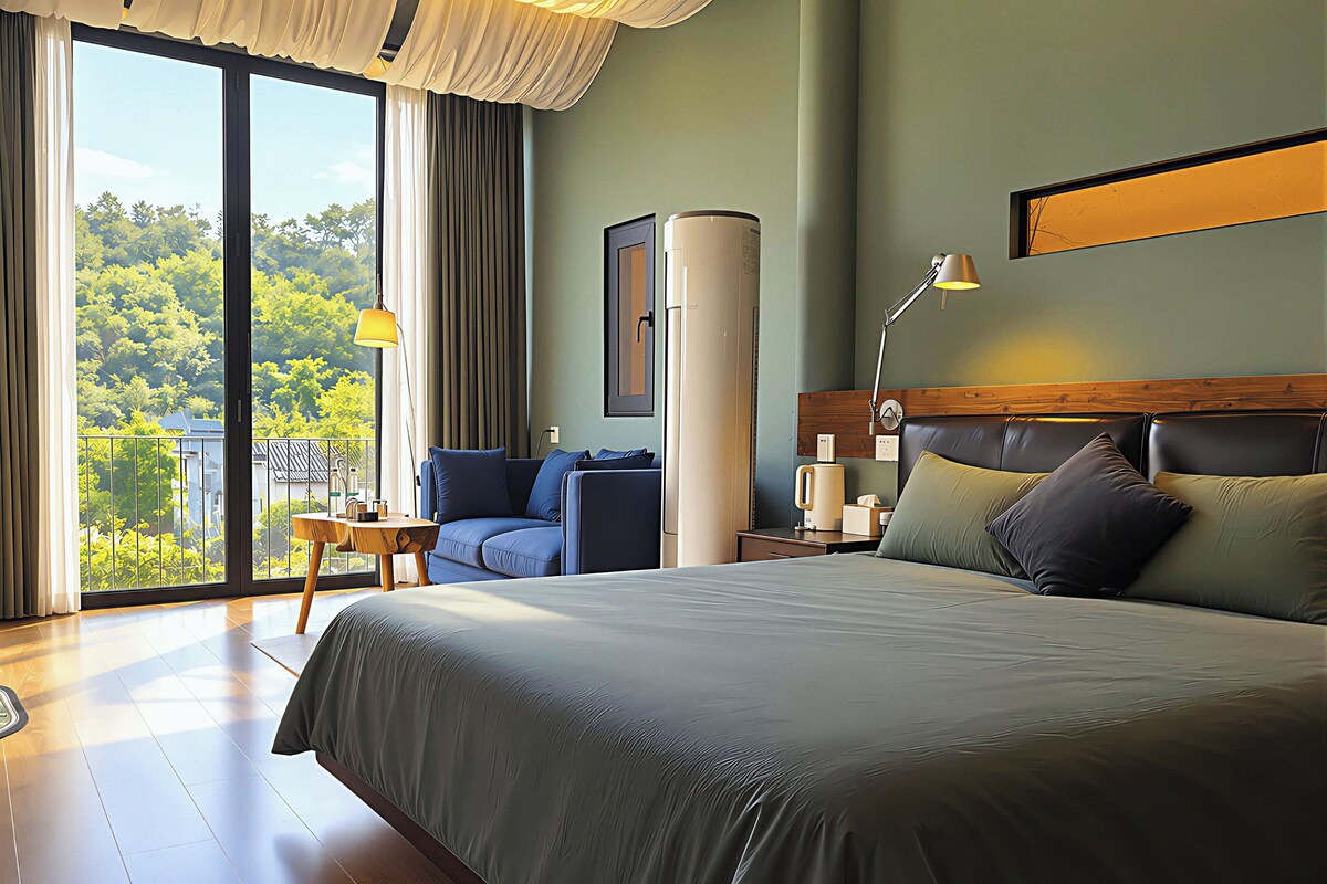 A hotel room with a large bed, a seating area with a table, and a view of greenery through the window.