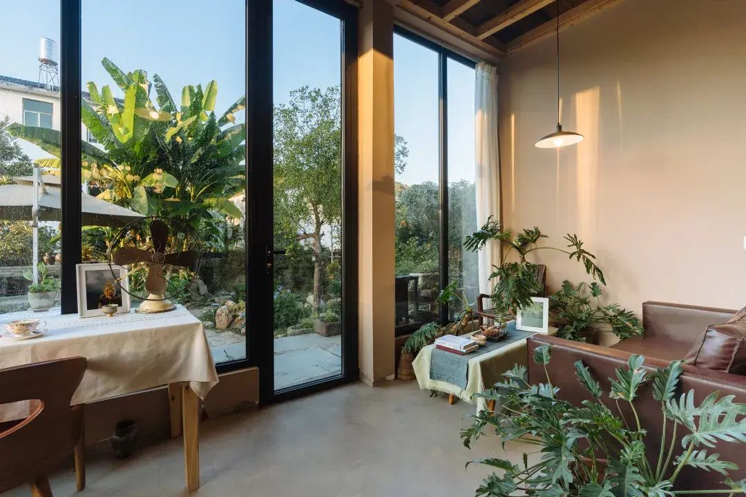 Una habitación con un ventanal grande, una mesa con mantel blanco, un sofá de cuero marrón y varias plantas en macetas.