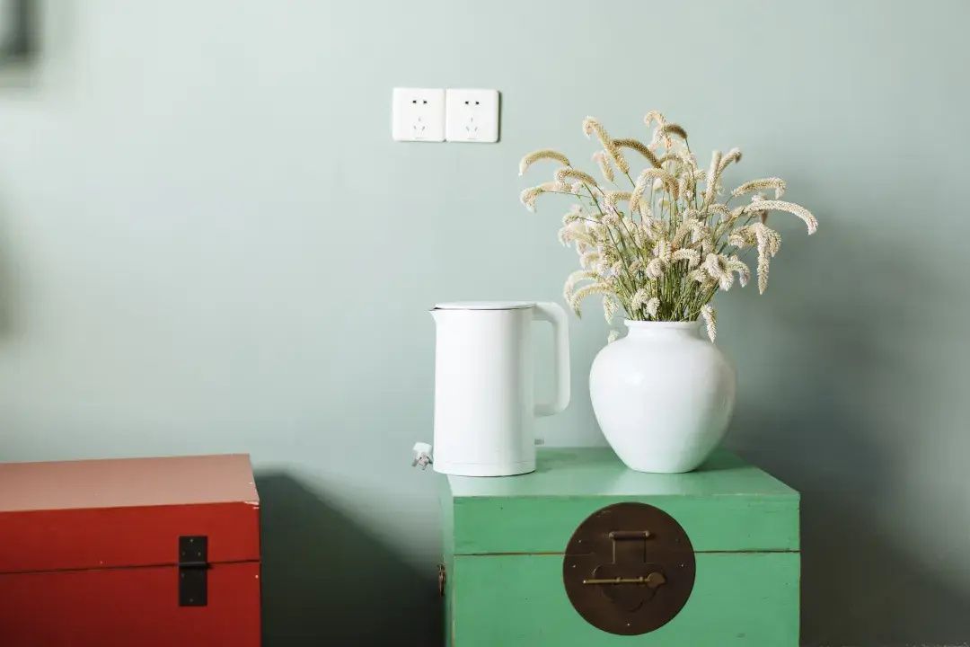 Uma cômoda verde com um vaso branco contendo flores secas e uma caneca branca em cima.
