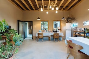 Un comedor con una mesa, sillas y lámparas colgantes. Hay plantas y una estantería en la habitación.