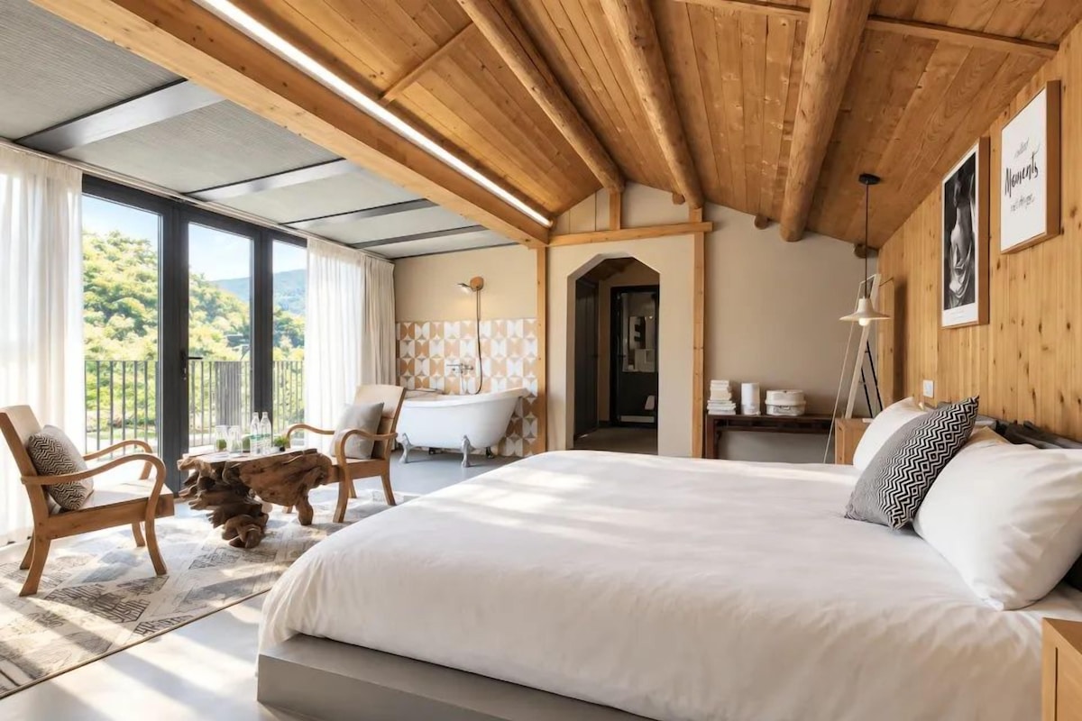 A spacious bedroom with a large bed, wooden ceiling beams, a view of greenery outside, and a bathroom with a bathtub and modern decor.