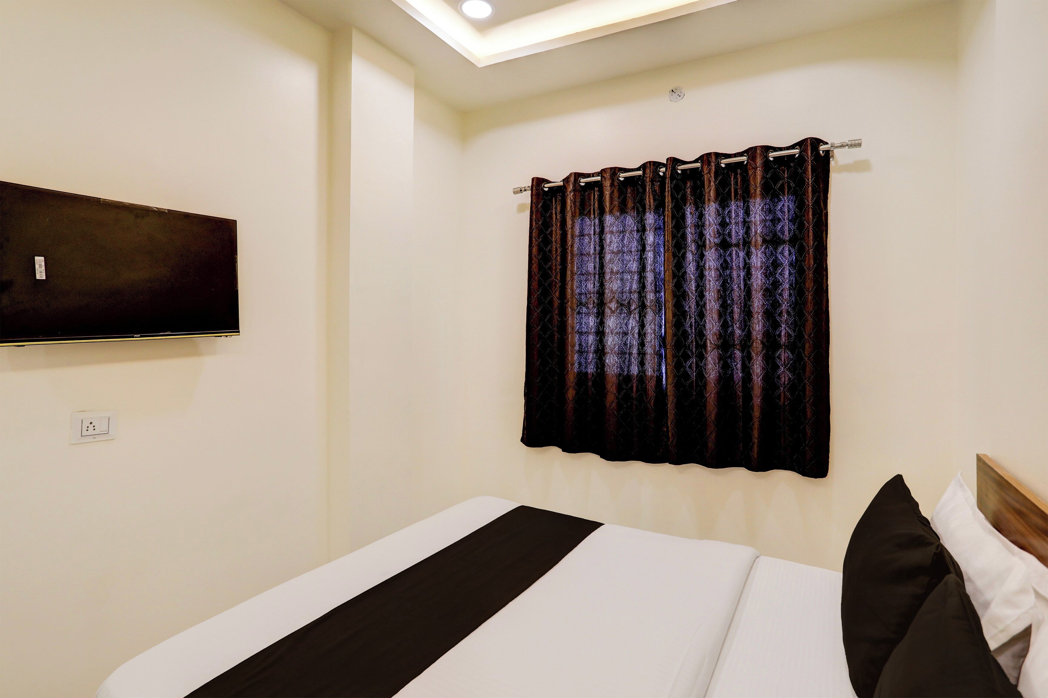 A bedroom with a bed, a flat-screen TV mounted on the wall, and a window with patterned curtains.