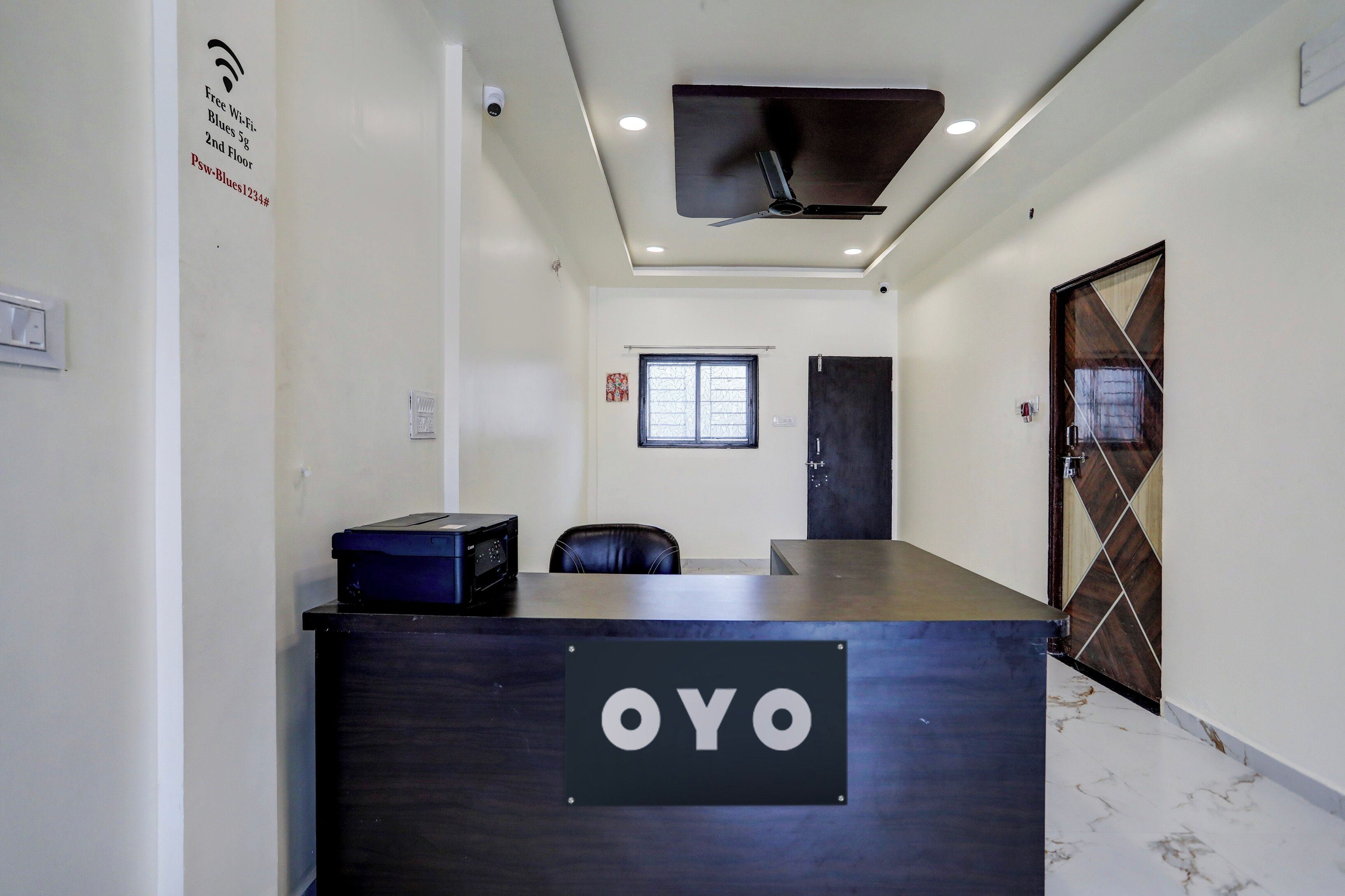 A reception desk with a sign reading 'OYO' in a hallway with a ceiling fan, a window, and a door.