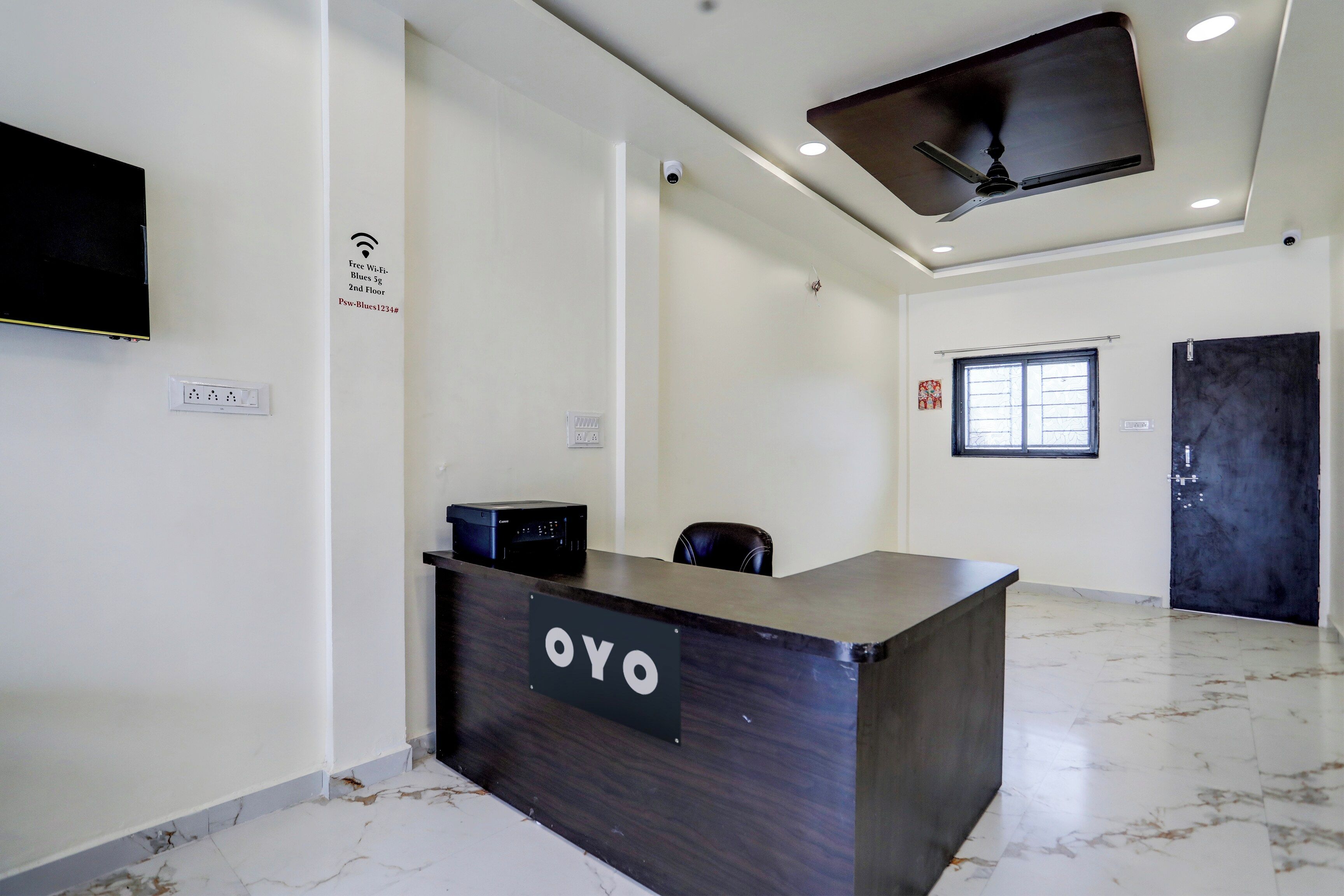 Un mostrador de recepción con un letrero que dice 'OYO' en el vestíbulo de un hotel, con un ventilador de techo, una ventana y una puerta.