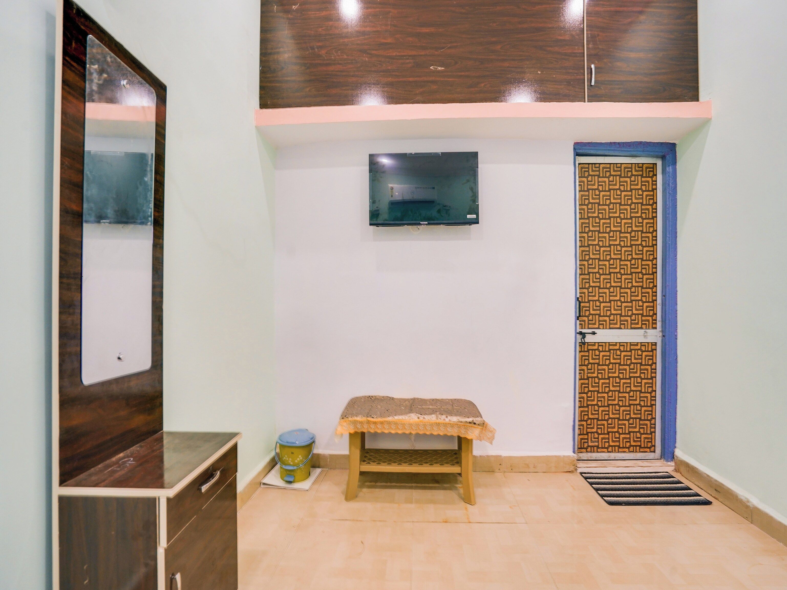 Quarto com um armário de madeira, um banco, uma televisão fixada na parede e uma porta estampada.