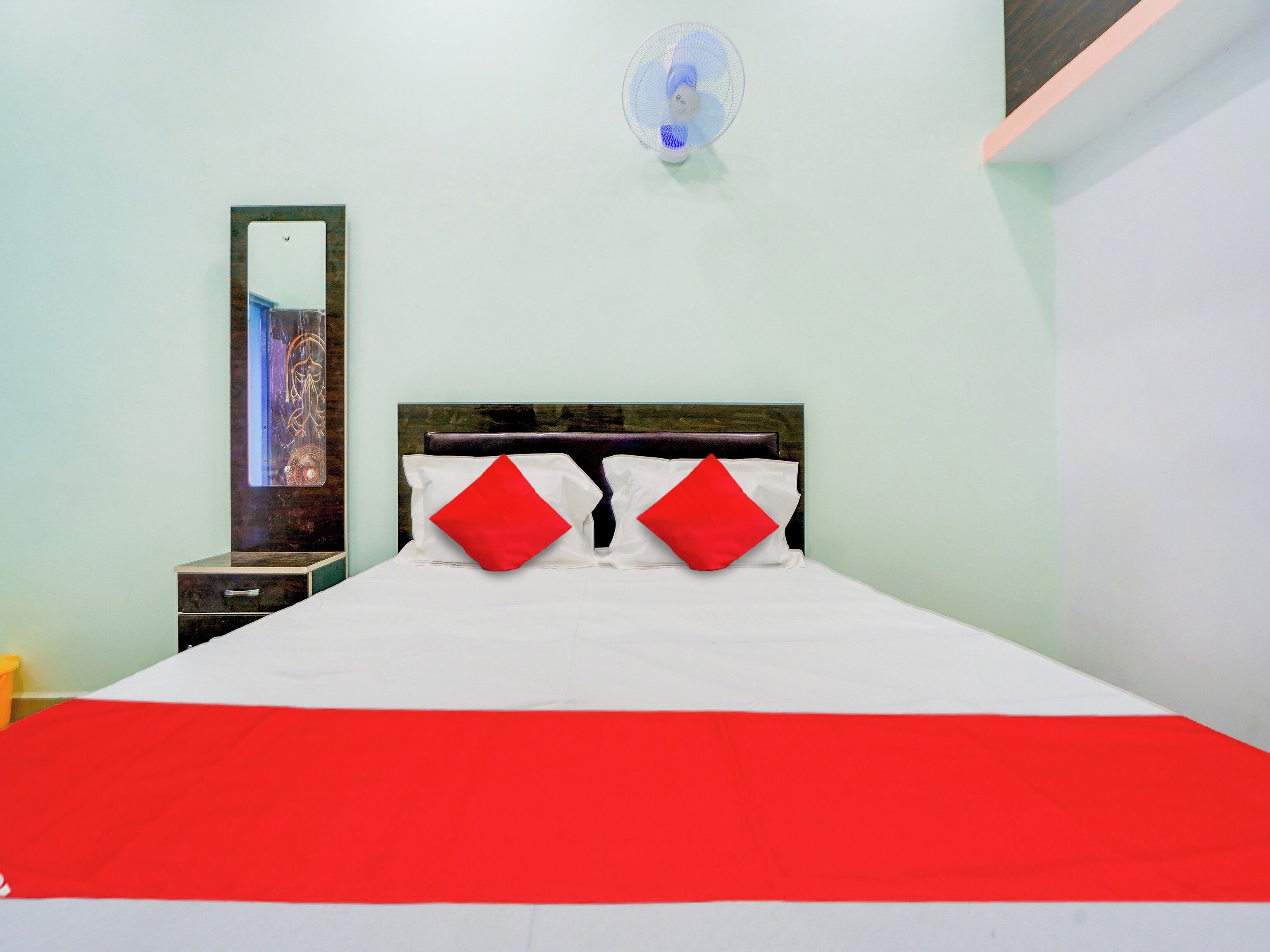 Quarto de hotel com cama, mesa de cabeceira, espelho e ventoinha fixada na parede.