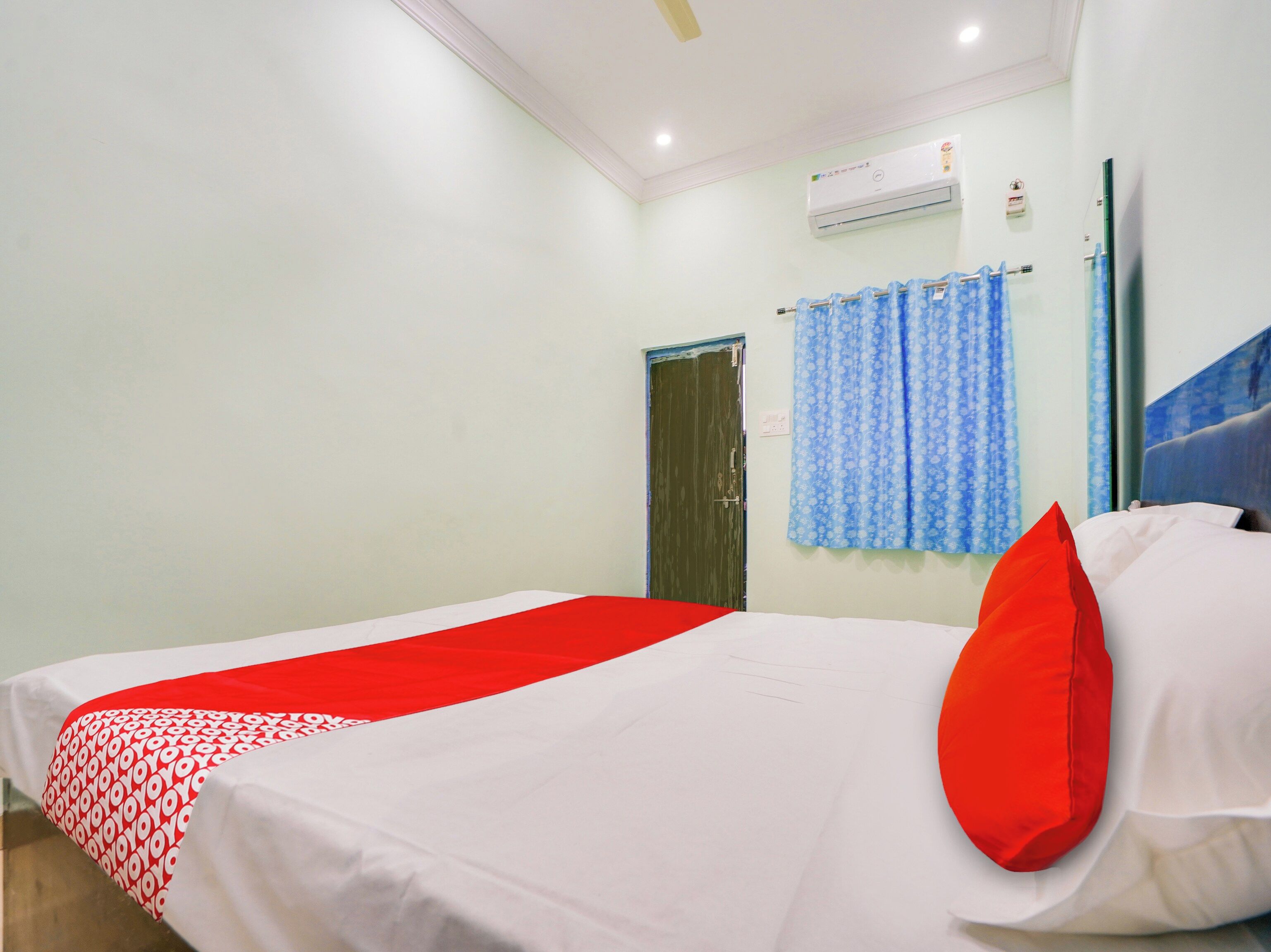 Una habitación de hotel con una cama, un almohadón rojo, una cortina azul y un equipo de aire acondicionado.