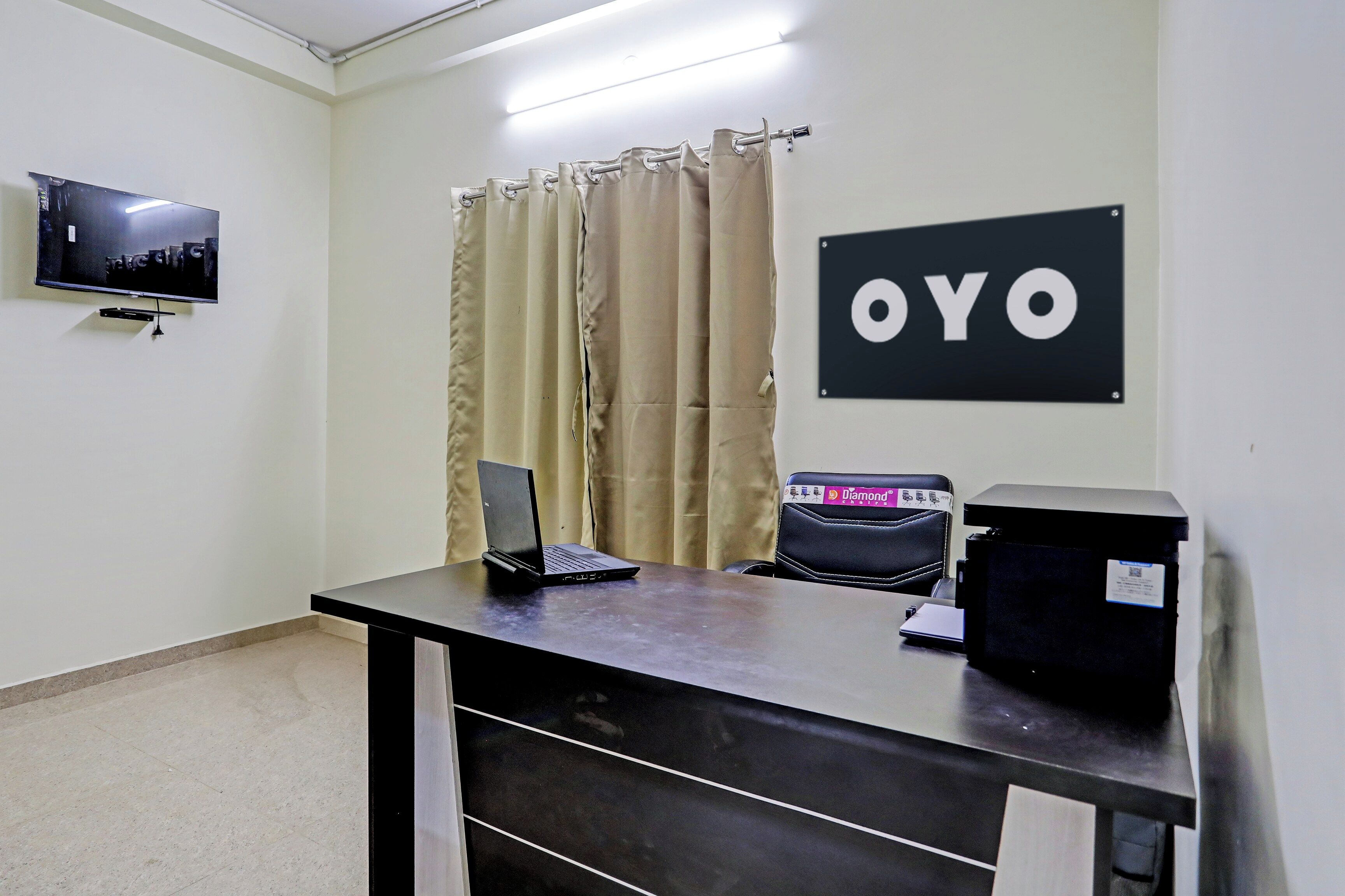 A reception desk with a laptop, a printer, and a chair. A television is mounted on the wall, and a sign with 'OYO' is visible.