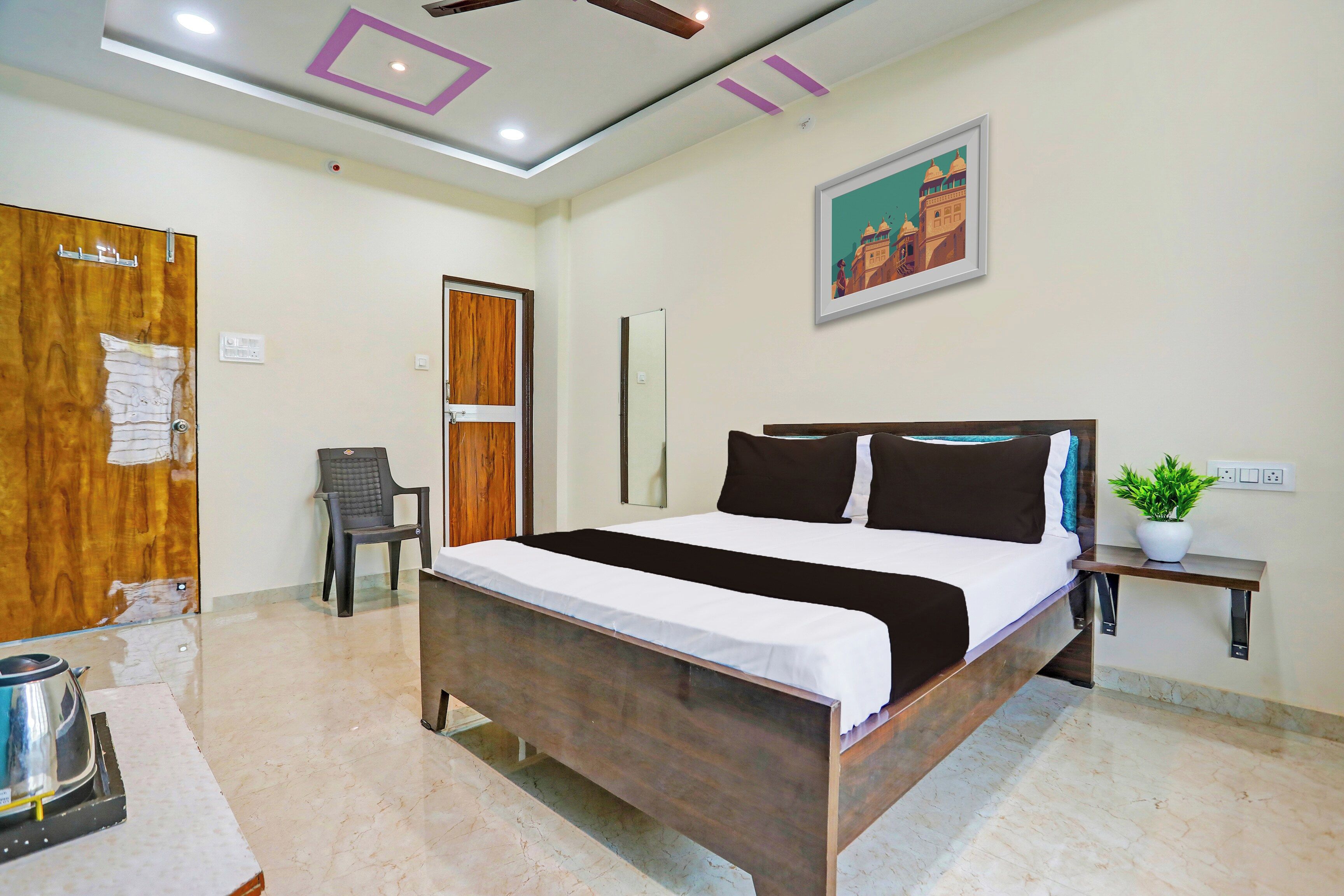 A hotel room with a bed, a chair, a bedside table, a wall-mounted picture, and a potted plant.