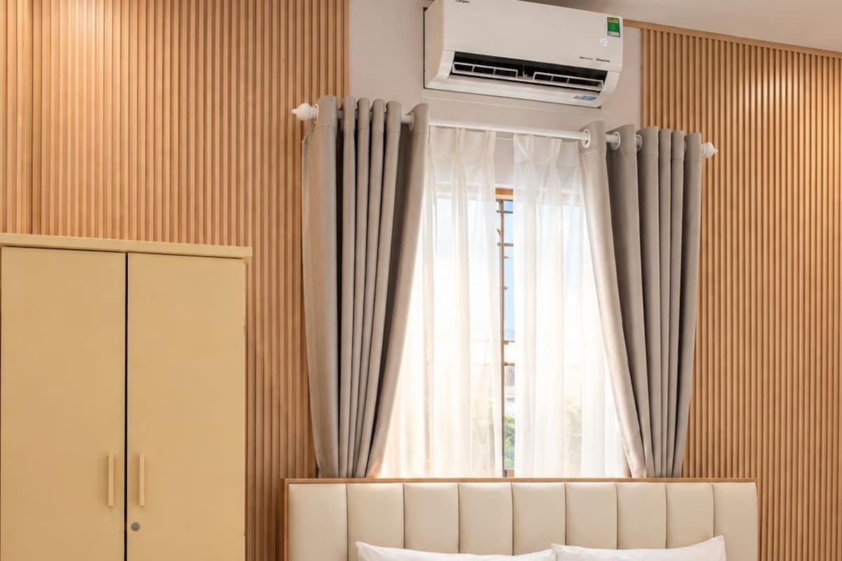 Una habitación de hotel con una cama, un armario, una ventana con cortinas y un equipo de aire acondicionado.